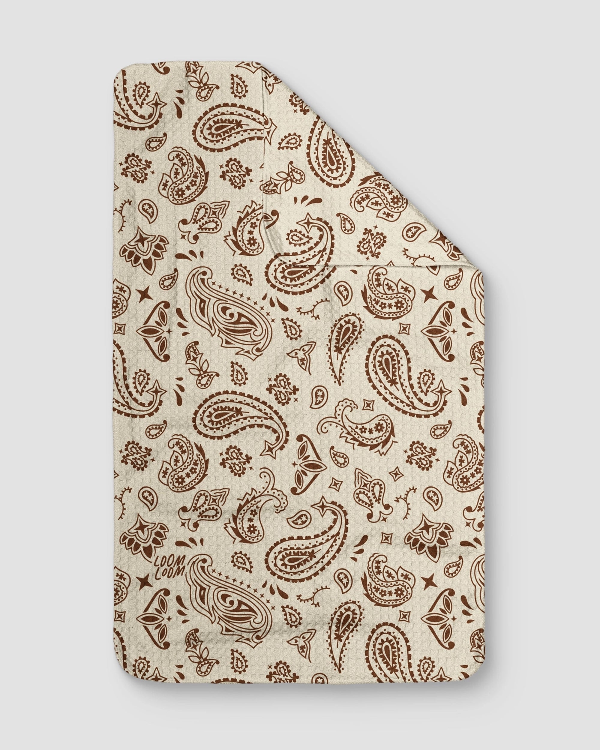 The Tan Bandana Towel by Faire is cream-colored with a brown paisley and floral pattern on super absorbent EcoLoom fabric. One corner is folded over to show the double-sided design and a small brown "Loom" logo in the lower left corner.