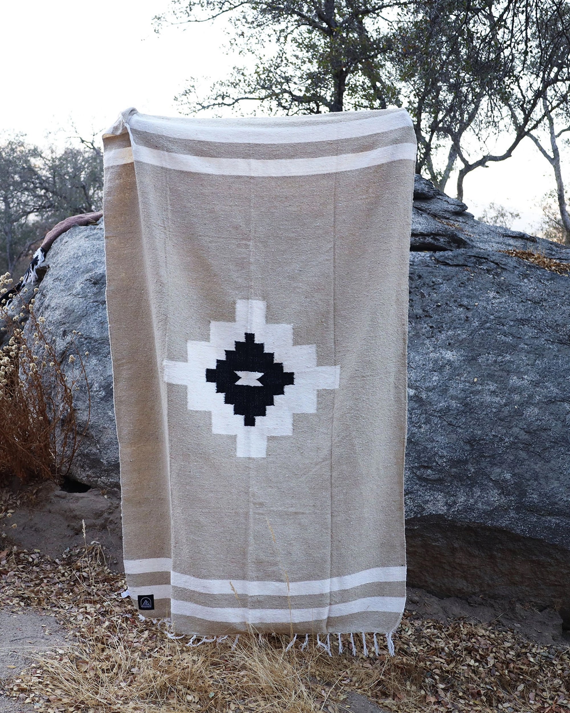 The TT Handwoven Blanket Taos Two by Faire, featuring a beige, white, and black geometric diamond pattern, is draped over a large gray rock outdoors among dry grass and trees.