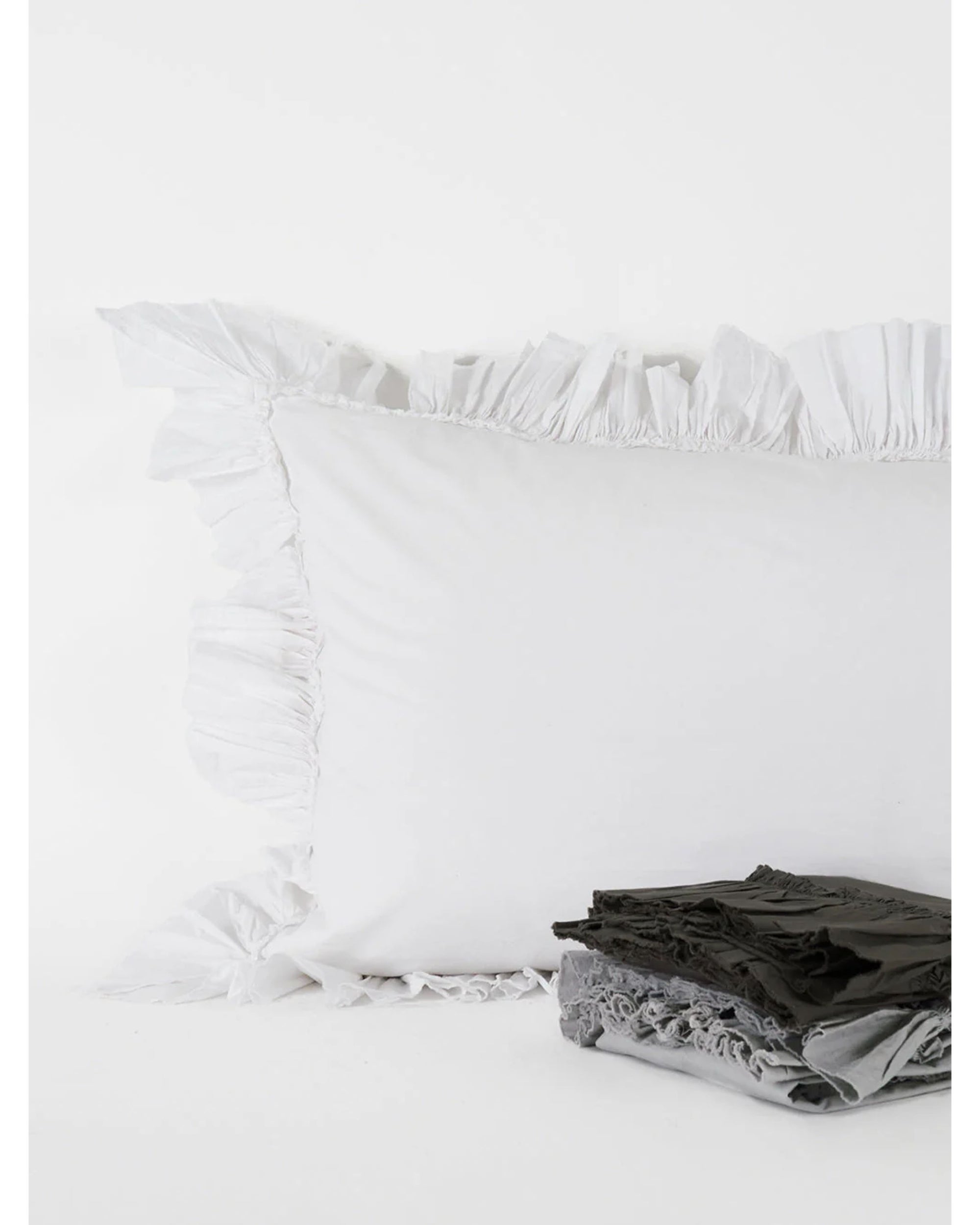 The Matteo Tat Cotton Sham in white, featuring a delicate ruffle border, rests against a white background. In front, gray and black textiles are neatly folded.