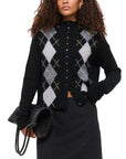 A woman with curly hair models the Alex Mill Taylor Cardigan Argyle, featuring a relaxed fit and black, gray, and white argyle pattern. She holds a textured black clutch and pairs it with a dark skirt against a plain background.