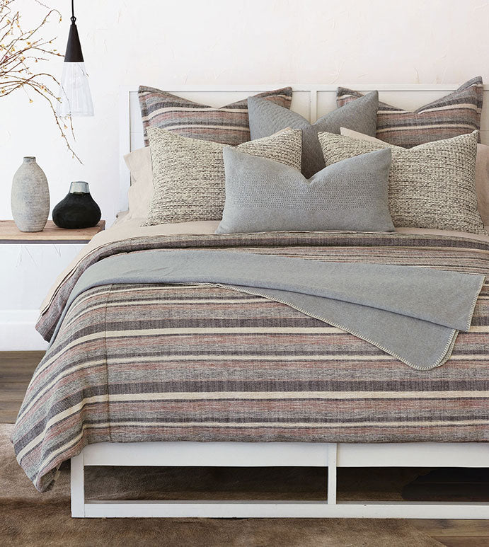 A neatly made bed with Ridge Striped 27x27" bedding in shades of beige, grey, and brown, adorned with a mix of textured pillows and a grey throw blanket. A minimalist lamp and vases decorate the background of this Scottsdale Arizona bungalow by Eastern Accents.