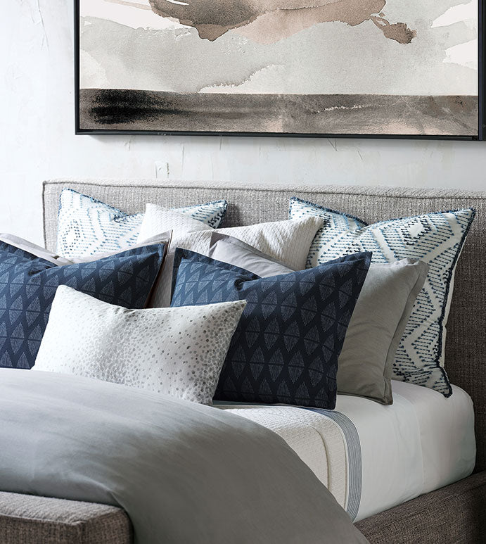 A neatly made bed features blue, gray, and white pillows, including the Eastern Accents BRIDGEHAMPTON STANDARD. Above the upholstered headboard on a light wall hangs a large abstract painting in neutral tones.