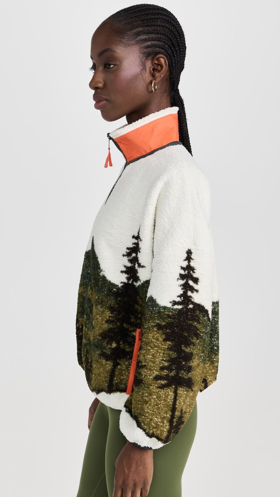 A woman with cornrowed hair wears The Plush Terrain Full-Zip by The Great Inc., a fleece jacket with a forest landscape pattern, orange-trimmed collar, and zip pockets, paired with green leggings against a light background.