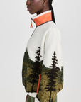 A woman with cornrowed hair wears The Plush Terrain Full-Zip by The Great Inc., a fleece jacket with a forest landscape pattern, orange-trimmed collar, and zip pockets, paired with green leggings against a light background.