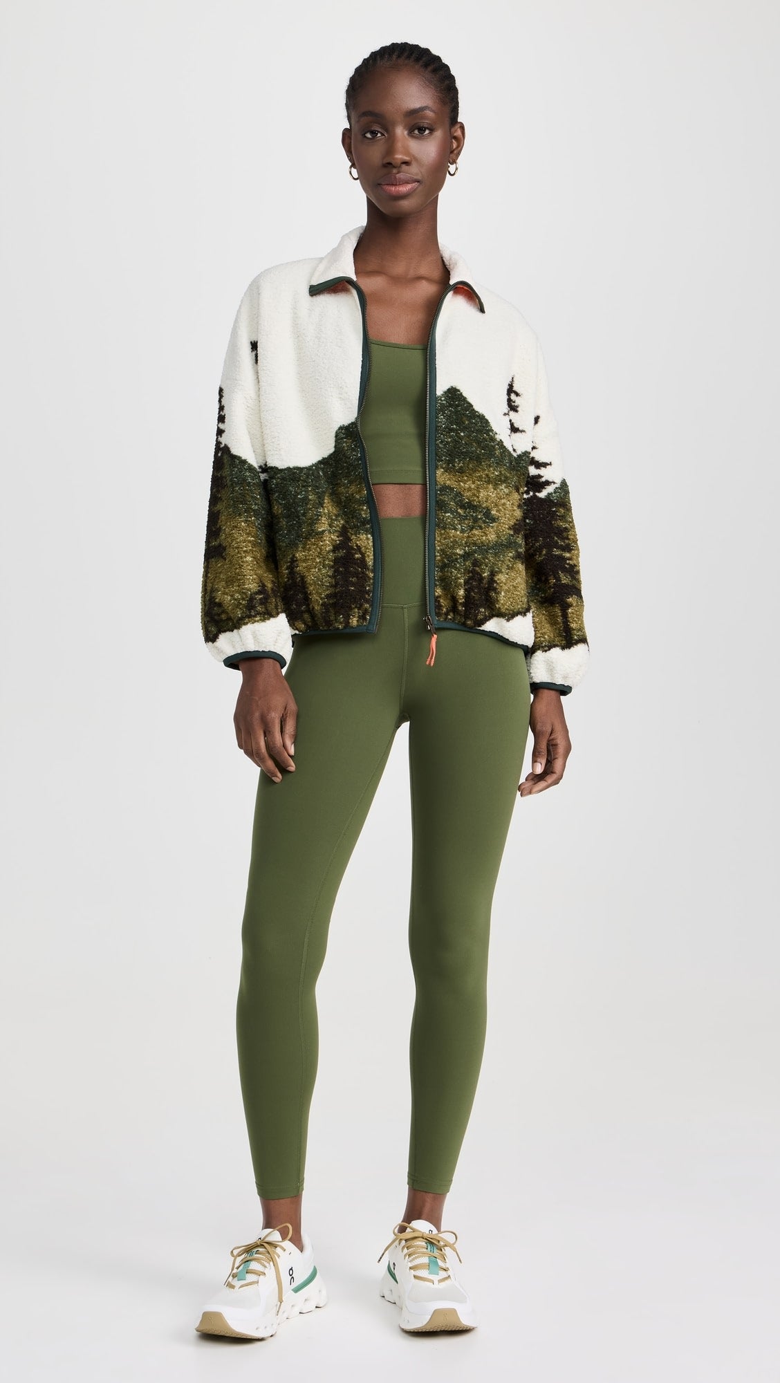A woman wears The Great Inc.'s The Plush Terrain Full-Zip—a white fleece jacket with a landscape pattern—over a green sports bra and leggings, paired with white and green sneakers, standing against a plain white background.