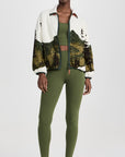 A woman wears The Great Inc.'s The Plush Terrain Full-Zip—a white fleece jacket with a landscape pattern—over a green sports bra and leggings, paired with white and green sneakers, standing against a plain white background.