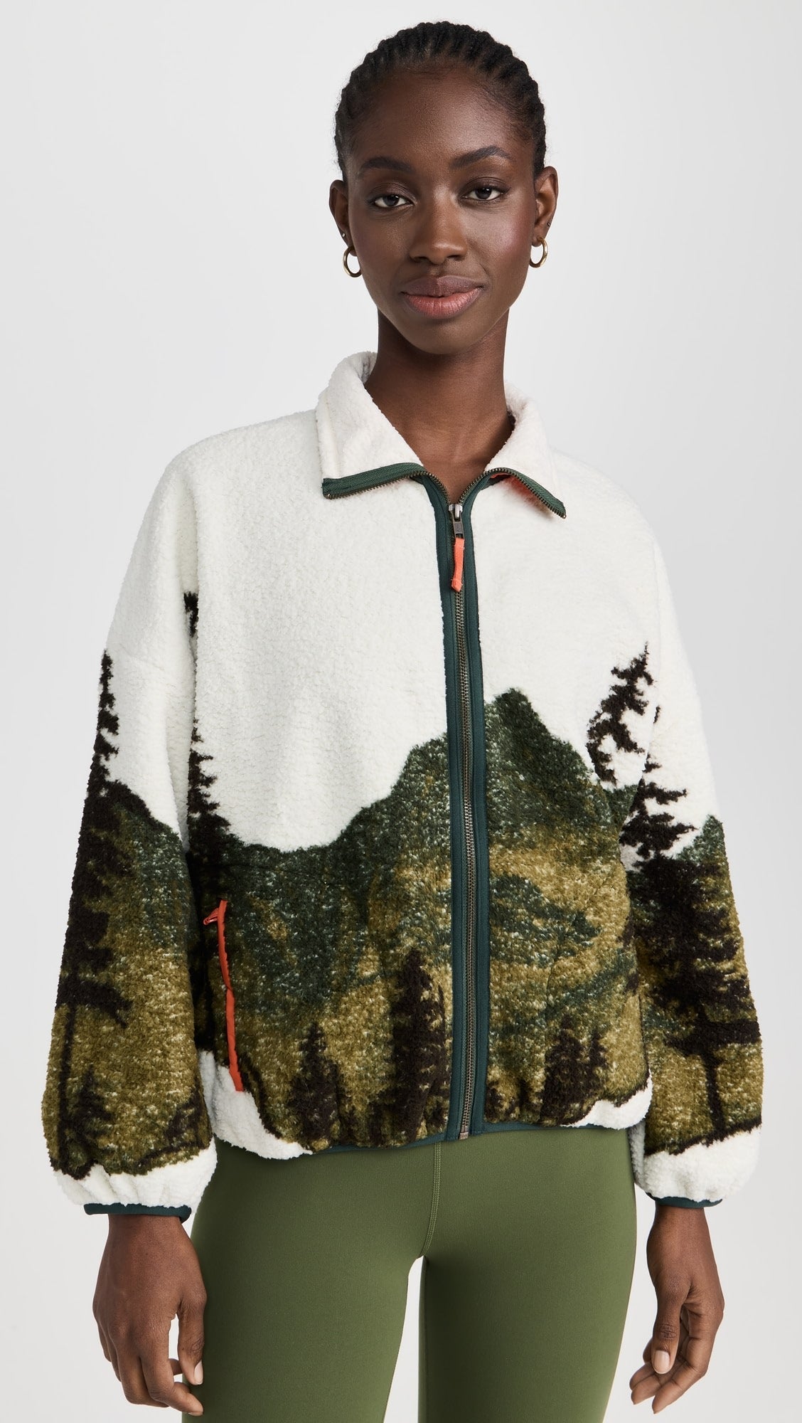 A woman wears The Great Inc.'s The Plush Terrain Full-Zip fleece jacket, featuring a scenic landscape print, zip front pockets, green zipper accents, and green leggings, standing against a plain white background.