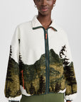 A woman wears The Great Inc.'s The Plush Terrain Full-Zip fleece jacket, featuring a scenic landscape print, zip front pockets, green zipper accents, and green leggings, standing against a plain white background.