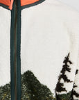 The Great Inc. Plush Terrain Full-Zip features a plush fleece build, zip front pockets, forest green zipper, green mountain and tree landscape pattern, and an orange collar with green trim and a zipper pull.