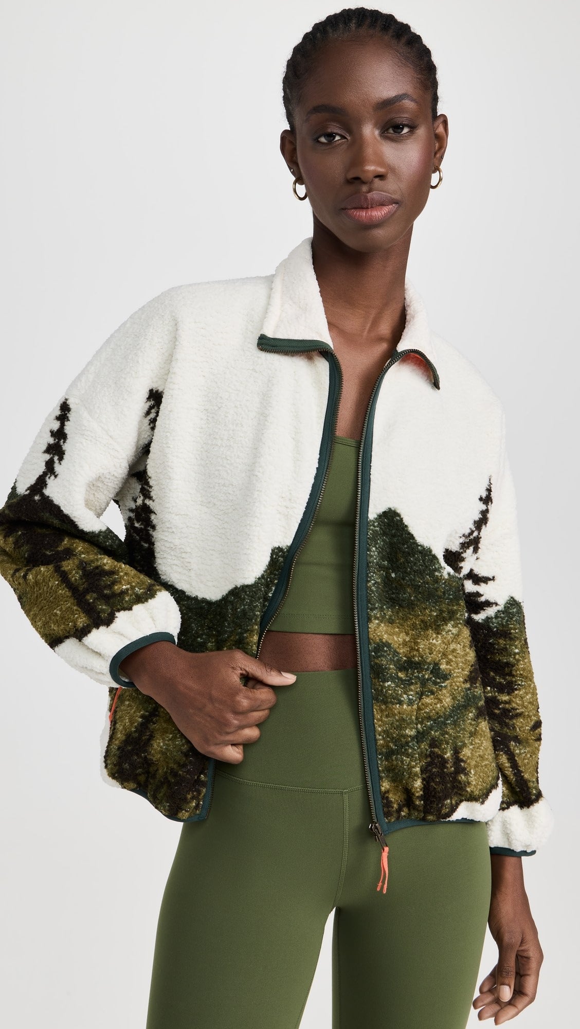 A woman in The Great Inc. Plush Terrain Full-Zip, featuring a mountain landscape design, stands facing the camera with her hand partly opening the zippered front pockets.