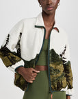 A woman in The Great Inc. Plush Terrain Full-Zip, featuring a mountain landscape design, stands facing the camera with her hand partly opening the zippered front pockets.