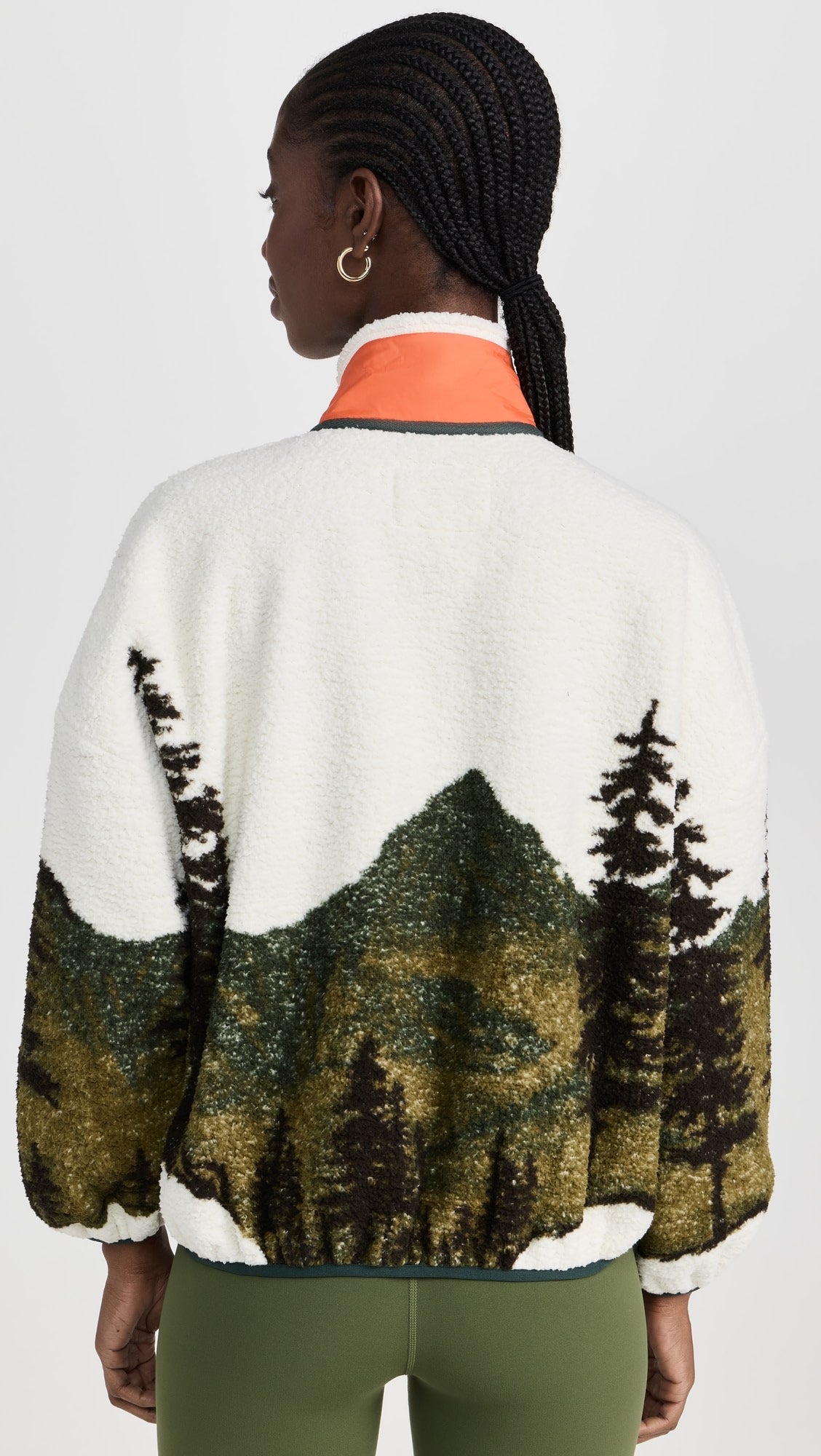 A person with braided hair is seen from behind wearing The Great Inc.’s Plush Terrain Full-Zip, a plush fleece jacket with a green mountain and tree landscape design and an orange collar.
