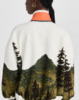 A person with braided hair is seen from behind wearing The Great Inc.’s Plush Terrain Full-Zip, a plush fleece jacket with a green mountain and tree landscape design and an orange collar.