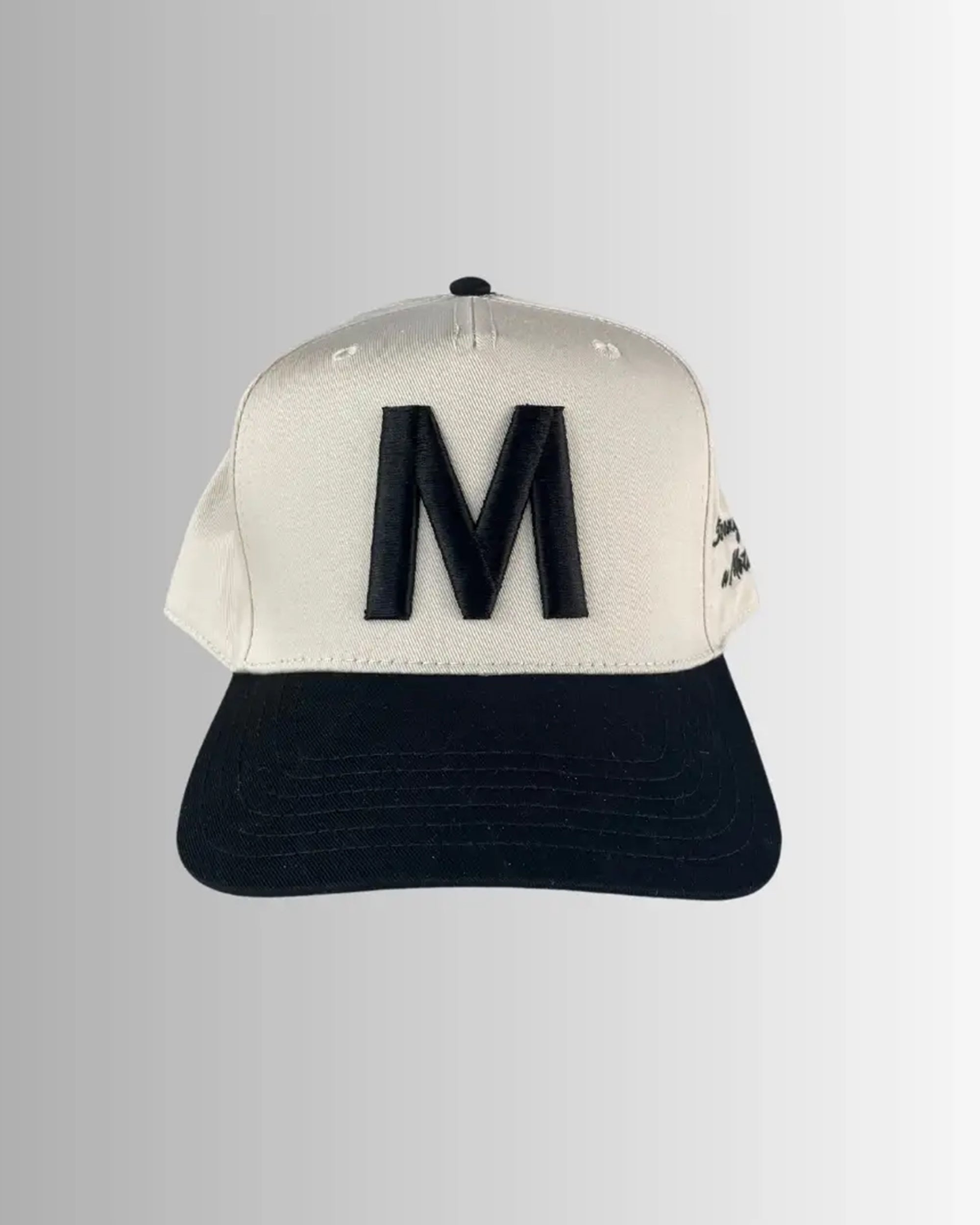 The M Hat by Faire features a white embroidered design with a bold black "M" on the front and a black brim, celebrating the "Strong as a mother" spirit. Shown against a gray gradient background.