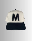 The M Hat by Faire features a white embroidered design with a bold black "M" on the front and a black brim, celebrating the "Strong as a mother" spirit. Shown against a gray gradient background.