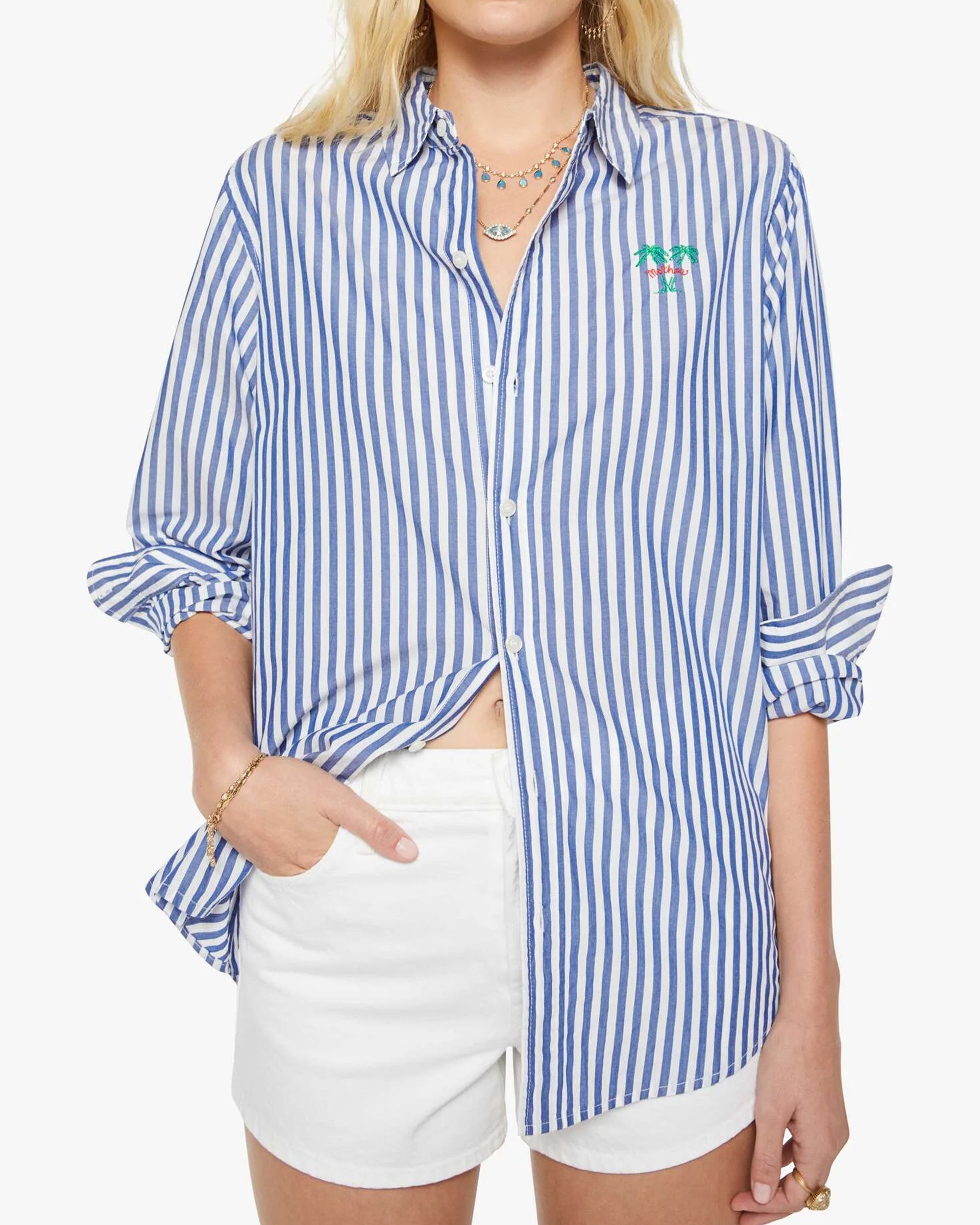 A person wears The Bestie by Mother—a blue and white striped cotton button-up shirt with rolled sleeves, a small green and pink embroidered detail on the chest—and white shorts, with one hand in their pocket.