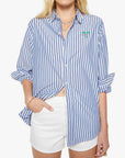 A person wears The Bestie by Mother—a blue and white striped cotton button-up shirt with rolled sleeves, a small green and pink embroidered detail on the chest—and white shorts, with one hand in their pocket.