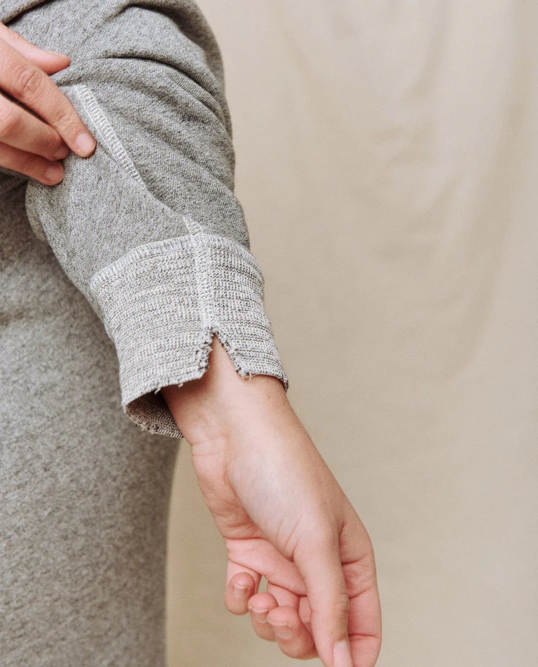 A person rolls up the sleeve of The Great Inc.'s College Sweatshirt in Varsity Grey, showcasing its textured cuff and small notch detail against a plain, light-colored fabric background.