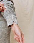 A person rolls up the sleeve of The Great Inc.'s College Sweatshirt in Varsity Grey, showcasing its textured cuff and small notch detail against a plain, light-colored fabric background.