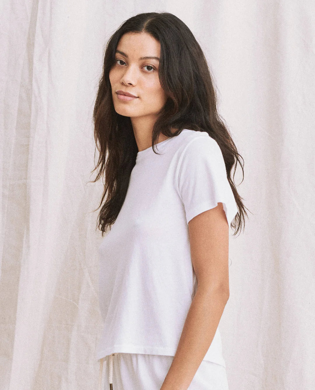 A woman with long dark hair wears The Little Tee by The Great Inc. in white Rancho Stripe, standing before a light fabric backdrop and gazing at the camera with a soft, neutral look.