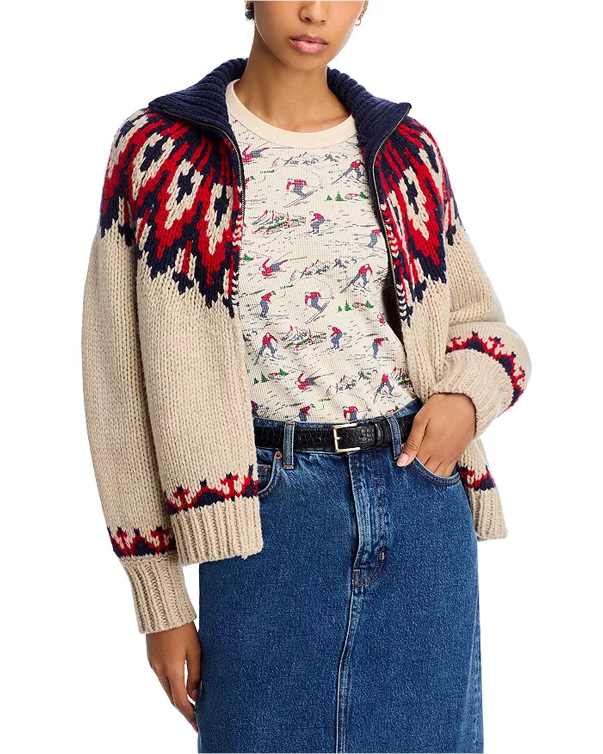 A person wears The Great Inc.’s Nordic Sweater in beige, red, and navy over a printed shirt with a blue denim skirt, one hand in their pocket. The look is casual and cozy.