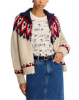 A person wears The Great Inc.’s Nordic Sweater in beige, red, and navy over a printed shirt with a blue denim skirt, one hand in their pocket. The look is casual and cozy.