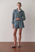 A person stands indoors against a plain light wall and wooden floor, wearing Donni's The Stripe Pop Boxer in blue-and-white stripes, paired with a light blue pullover, white t-shirt, and dark slide sandals.