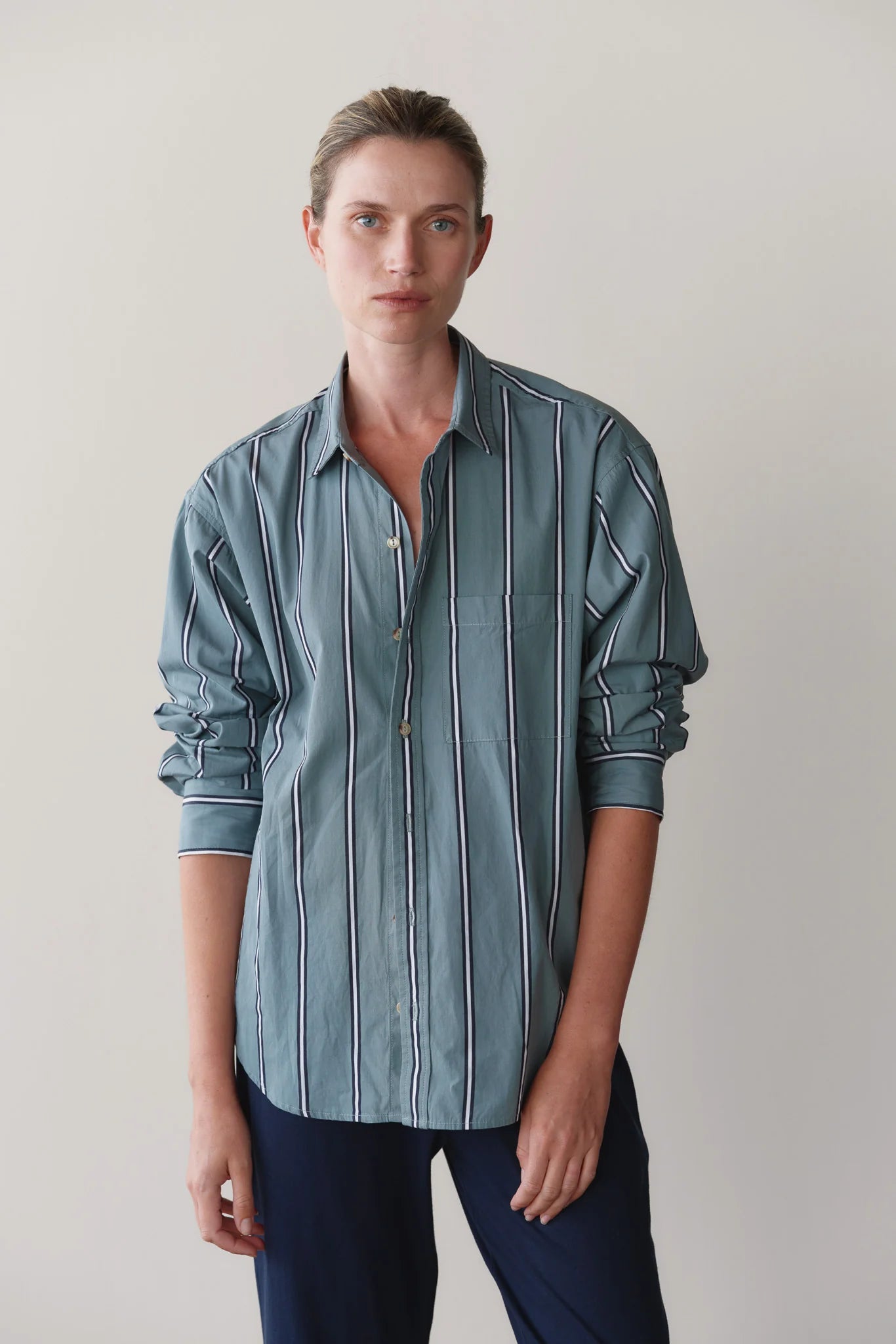 A person with short, light brown hair wears Donni's The Stripe Pop Shirt— a relaxed fit teal poplin top with white and dark stripes—paired with dark pants, standing against a plain light background.