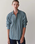 A person with short, light brown hair wears Donni's The Stripe Pop Shirt— a relaxed fit teal poplin top with white and dark stripes—paired with dark pants, standing against a plain light background.