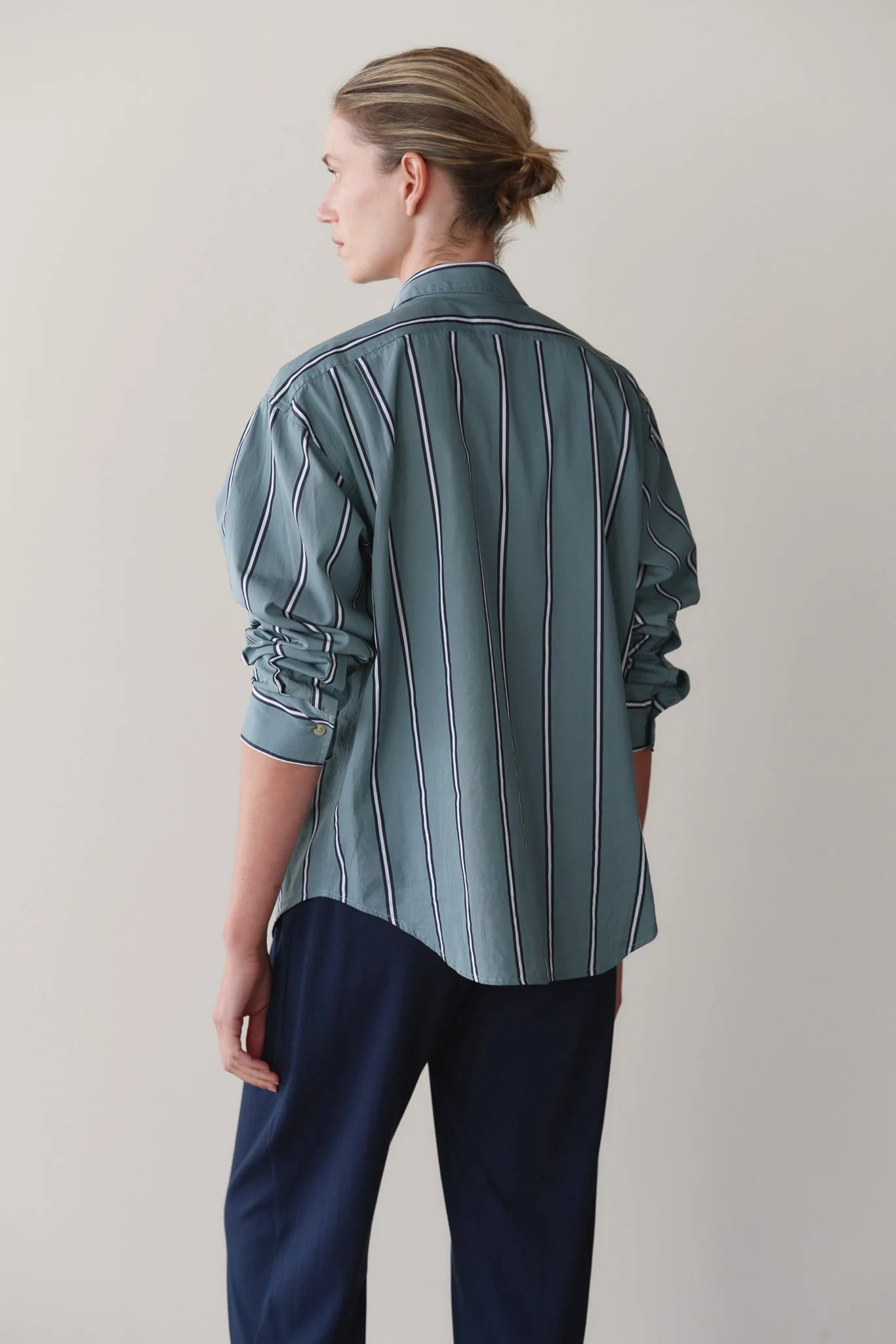 A person with light brown hair tied back is shown from behind, wearing Donni's The Stripe Pop Shirt in teal with vertical stripes and dark navy trousers against a plain light background.