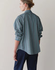 A person with light brown hair tied back is shown from behind, wearing Donni's The Stripe Pop Shirt in teal with vertical stripes and dark navy trousers against a plain light background.