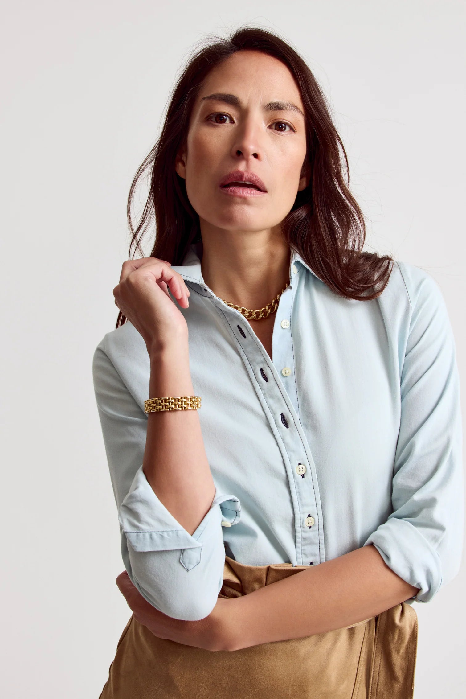 A person with long brown hair wears THE SHIRT’s Signature Shirt in light blue with No Gape button technology, styled with a gold bracelet and chain necklace, posing with one arm bent against a plain light background.