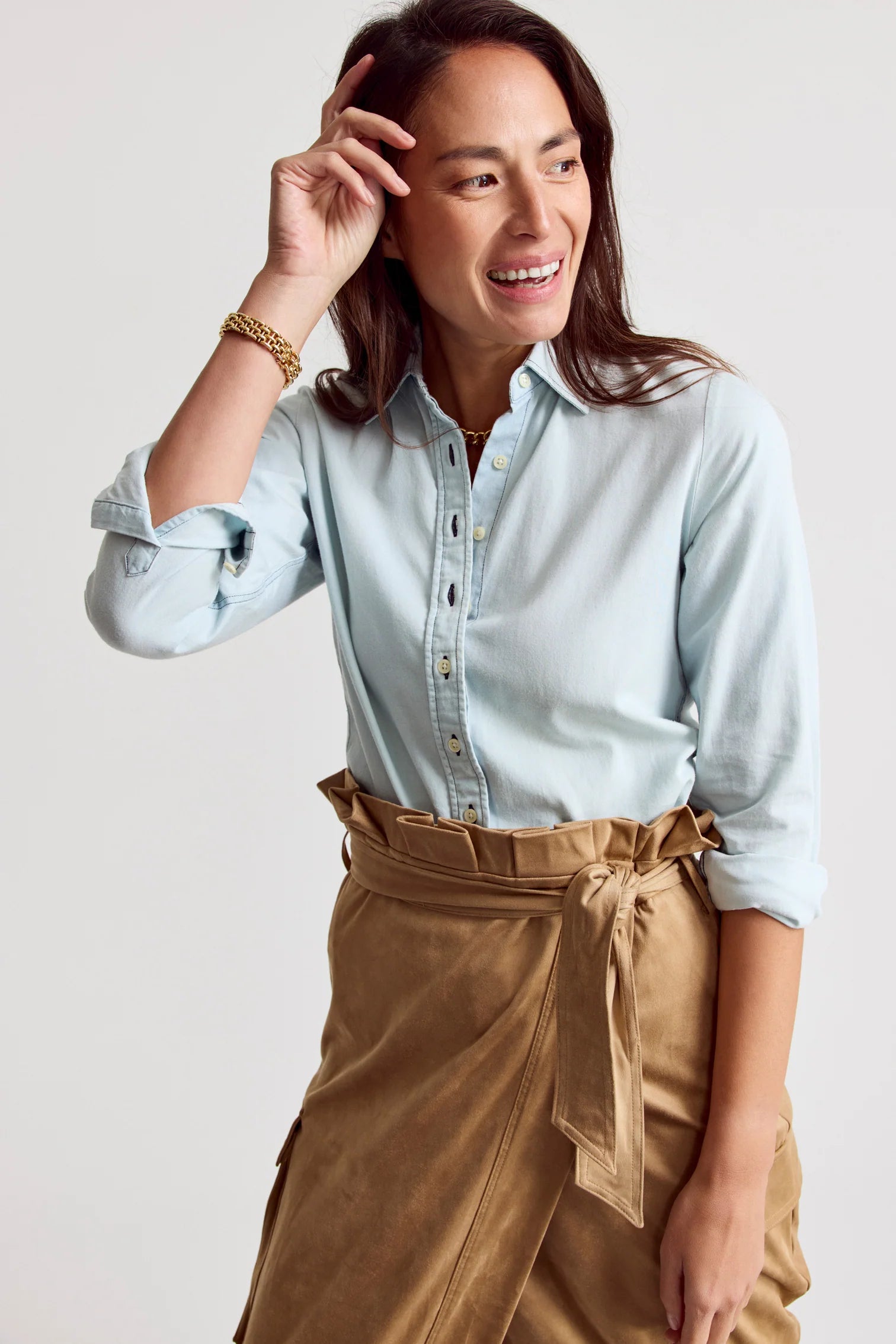 A woman smiles with her hand near her head, wearing THE SHIRT’s Signature Shirt in light blue with No Gape button technology and rolled-up sleeves, paired with a tan wrap skirt. The background is plain and light-colored.