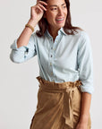 A woman smiles with her hand near her head, wearing THE SHIRT’s Signature Shirt in light blue with No Gape button technology and rolled-up sleeves, paired with a tan wrap skirt. The background is plain and light-colored.
