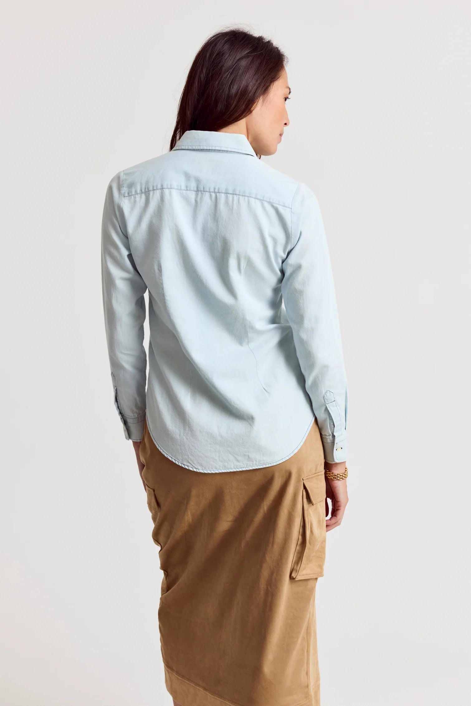 A woman with brown hair stands against a plain light background, wearing THE SHIRT’s Signature Shirt in light blue with No Gape button technology and a tan midi skirt featuring side pockets.