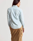 A woman with brown hair stands against a plain light background, wearing THE SHIRT’s Signature Shirt in light blue with No Gape button technology and a tan midi skirt featuring side pockets.