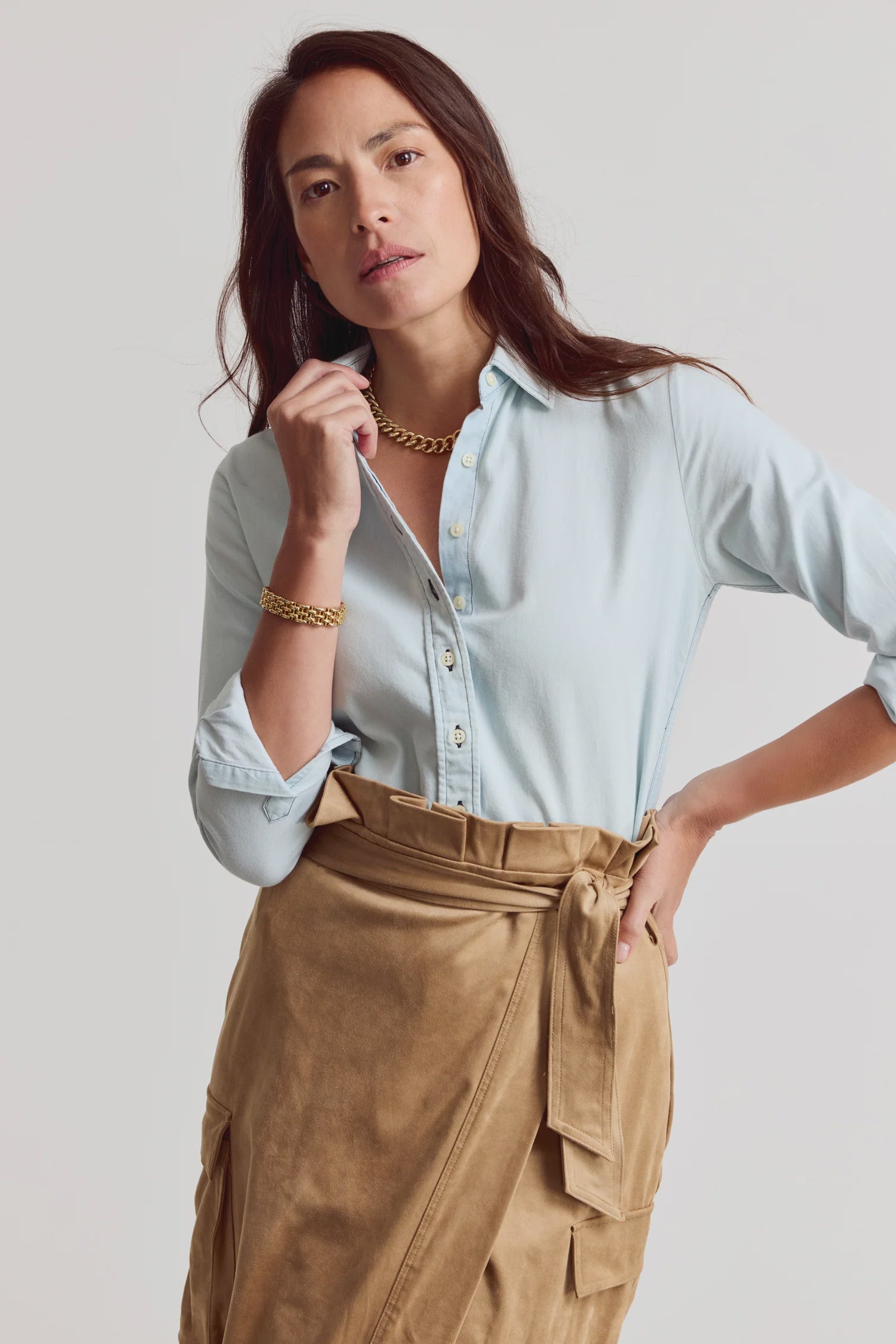 A woman poses against a light background in THE SHIRT's Signature Shirt in pale blue, featuring No Gape buttons and tucked into a tan wrap skirt. Gold jewelry completes her effortlessly chic look as she touches her collar.