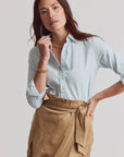 A woman poses against a light background in THE SHIRT's Signature Shirt in pale blue, featuring No Gape buttons and tucked into a tan wrap skirt. Gold jewelry completes her effortlessly chic look as she touches her collar.