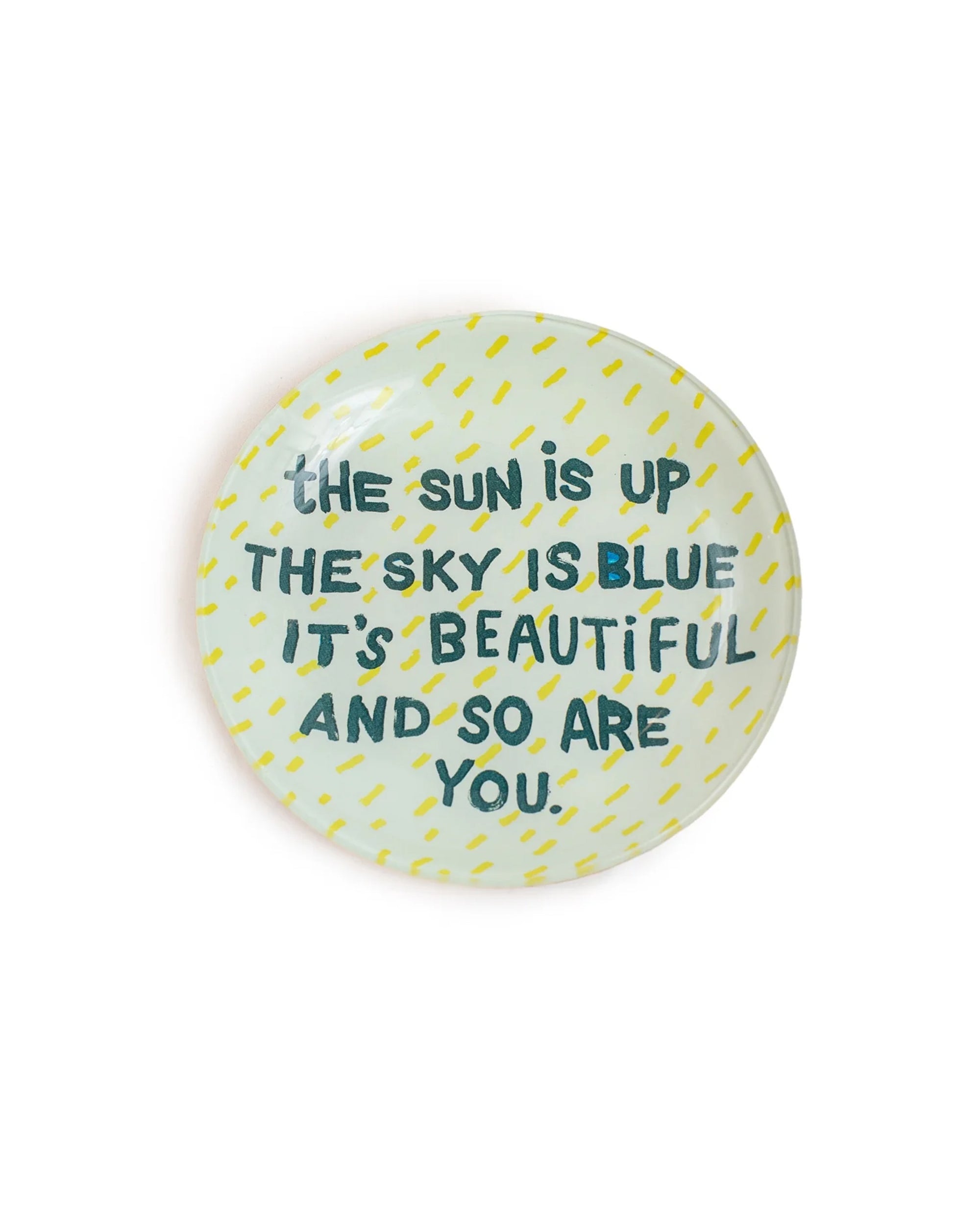The Sun Is Up Round Plate by Sugarboo Designs features a light blue background, yellow dashes, and the handwritten text "THE SUN IS UP THE SKY IS BLUE IT'S BEAUTIFUL AND SO ARE YOU"—ideal as a jewelry holder or decorative accent.