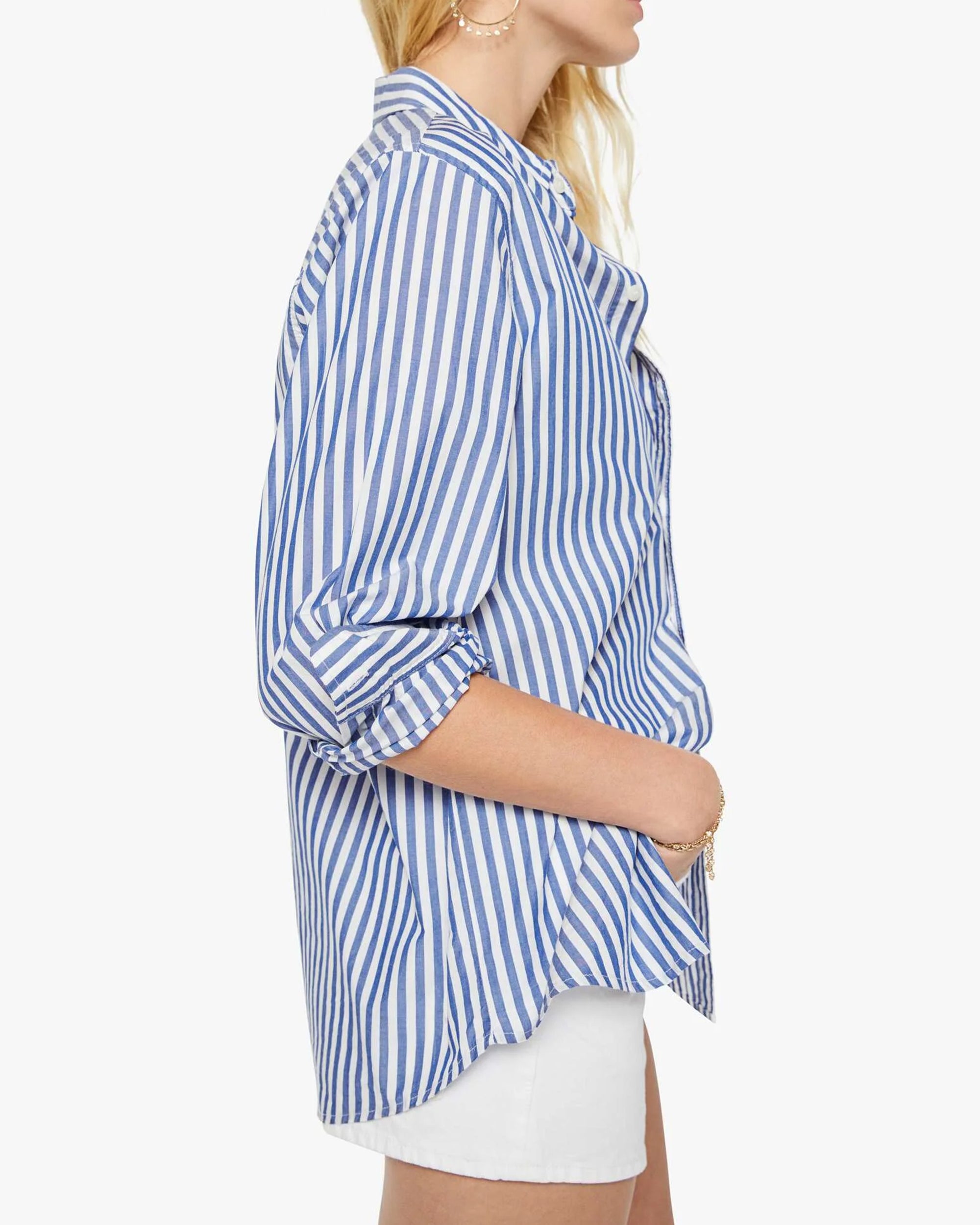A woman poses in profile against a white background, wearing The Bestie blue and white striped cotton button-up shirt with rolled sleeves from Mother, paired with white shorts.
