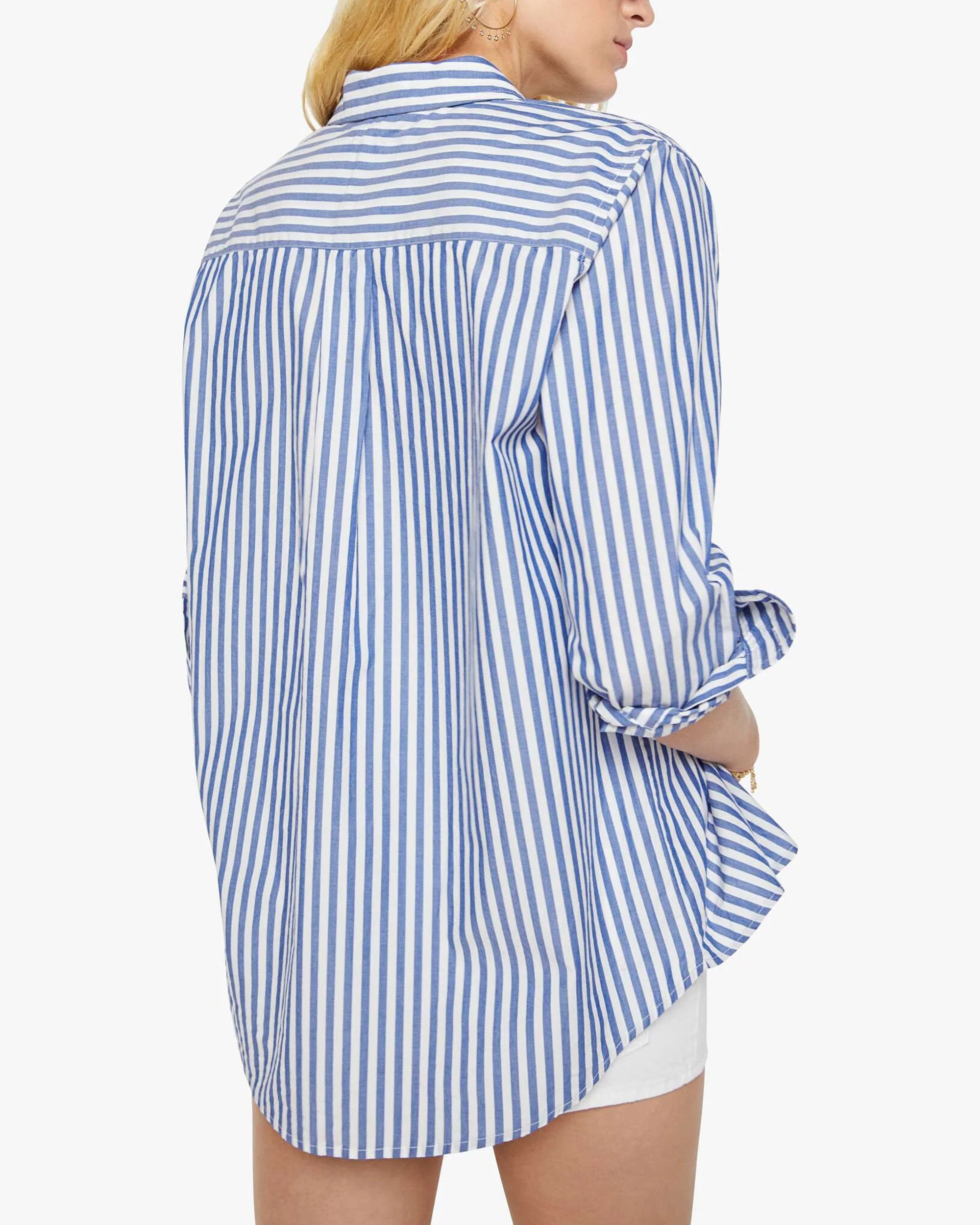 A person with long blonde hair is seen from the back wearing Mother's The Bestie—a loose-fitting 100% cotton blue and white striped button-up shirt with rolled sleeves—paired with white shorts.