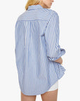 A person with long blonde hair is seen from the back wearing Mother's The Bestie—a loose-fitting 100% cotton blue and white striped button-up shirt with rolled sleeves—paired with white shorts.