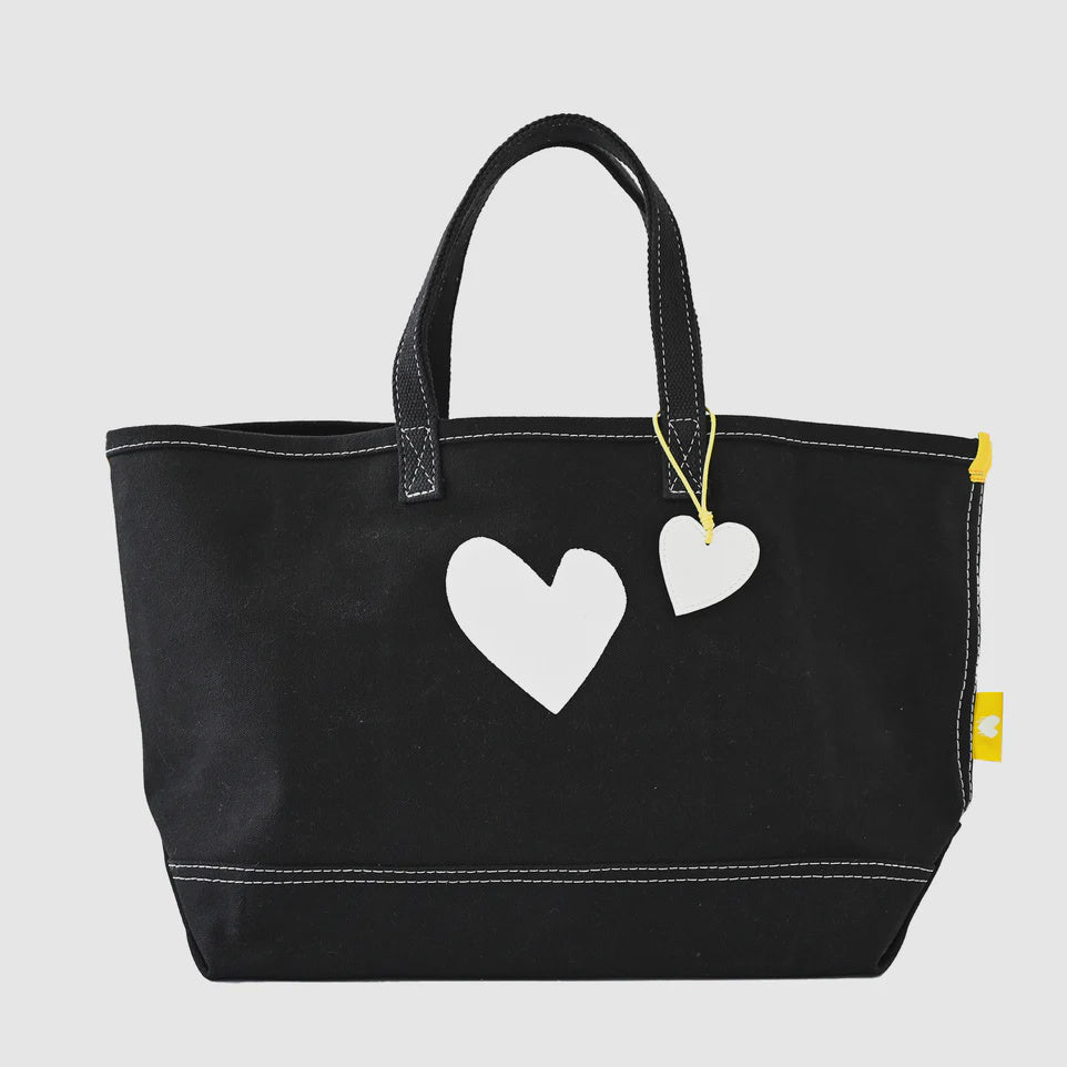 The Small Tote Imperfect Heart by Kerri Rosenthal is a black canvas tote bag with two white heart designs on the front. It has two short handles and a small white heart-shaped tag attached to one handle. Made from durable cotton canvas, it features white stitching and a small yellow tag on the right side.