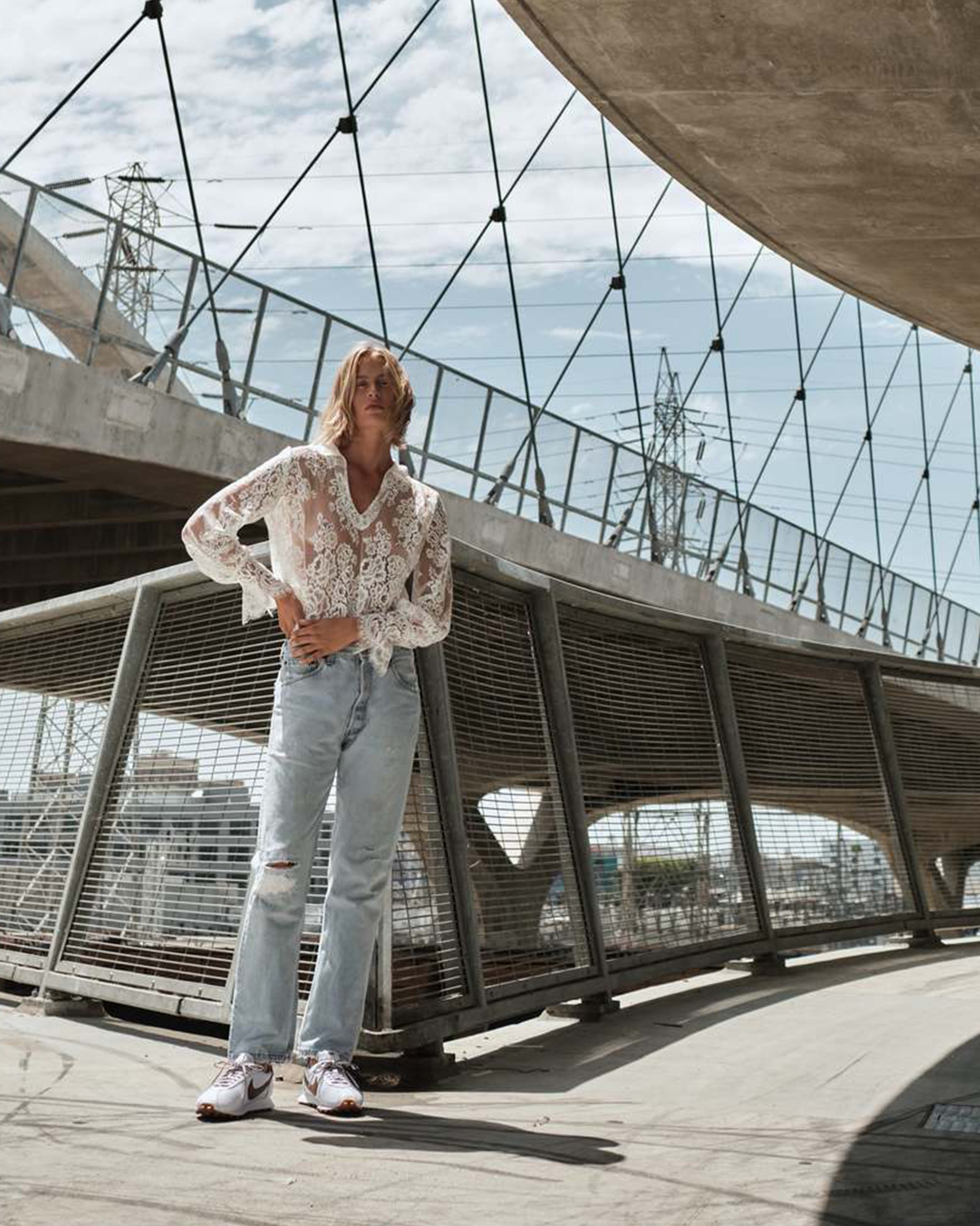 A person wearing The Victorian Shirt by Cissa, paired with light blue ripped jeans and white sneakers, stands outdoors under a modern bridge with cables and geometric structures on a partly cloudy day.