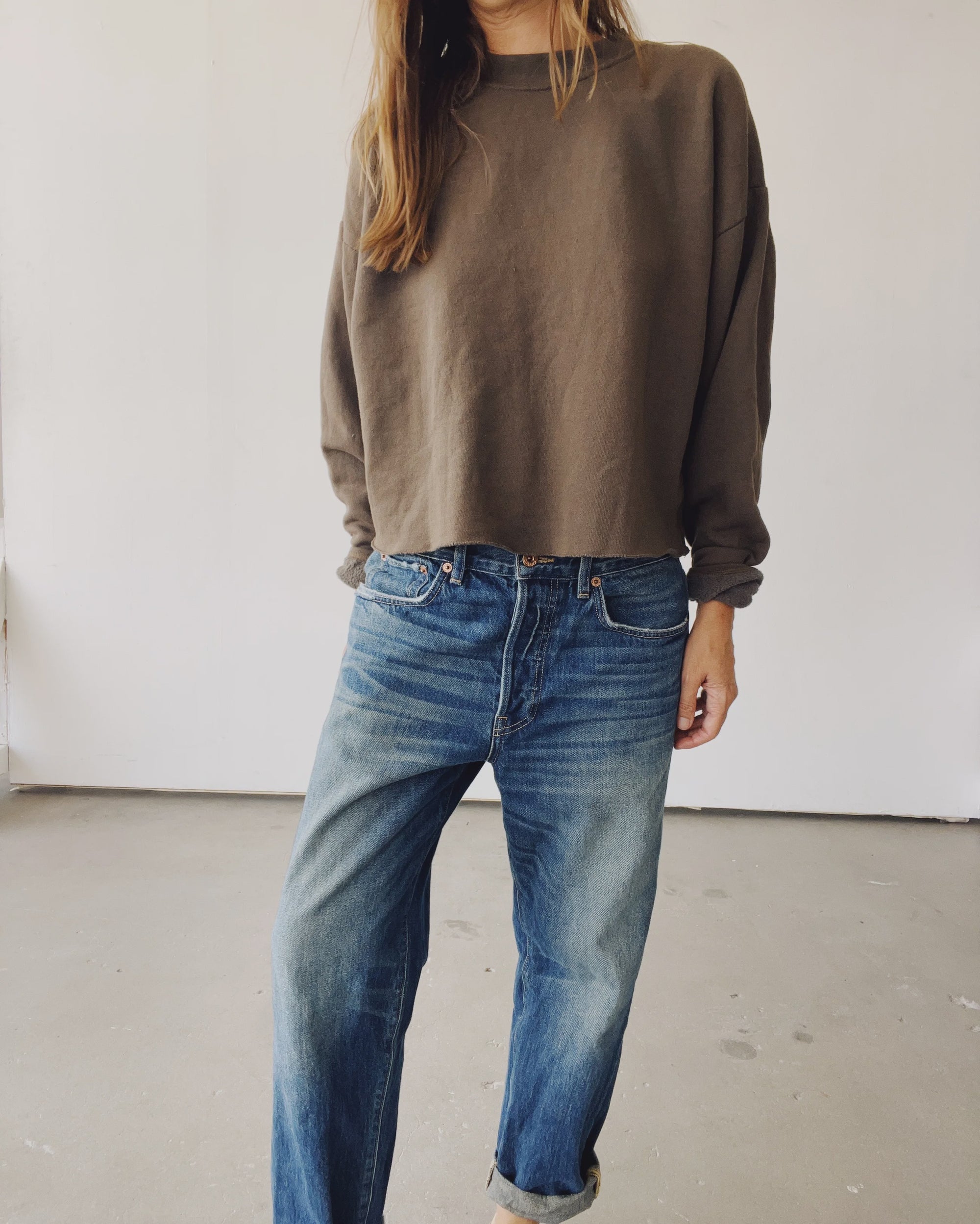Indoors, a person wears Hey Gang’s The Boxy Crop Sweatshirt in brown tri-blend fleece paired with relaxed blue jeans with rolled cuffs; their face remains unseen.