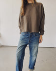 Indoors, a person wears Hey Gang’s The Boxy Crop Sweatshirt in brown tri-blend fleece paired with relaxed blue jeans with rolled cuffs; their face remains unseen.