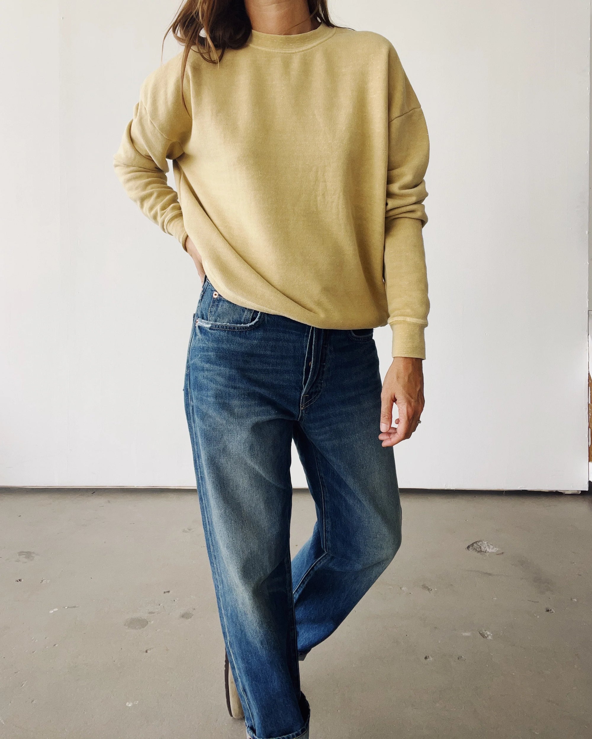 A person stands indoors against a white wall, wearing The Sweatshirt 26 by Hey Gang in beige with blue jeans. Their right hand rests on their hip and their face is partially out of the frame.