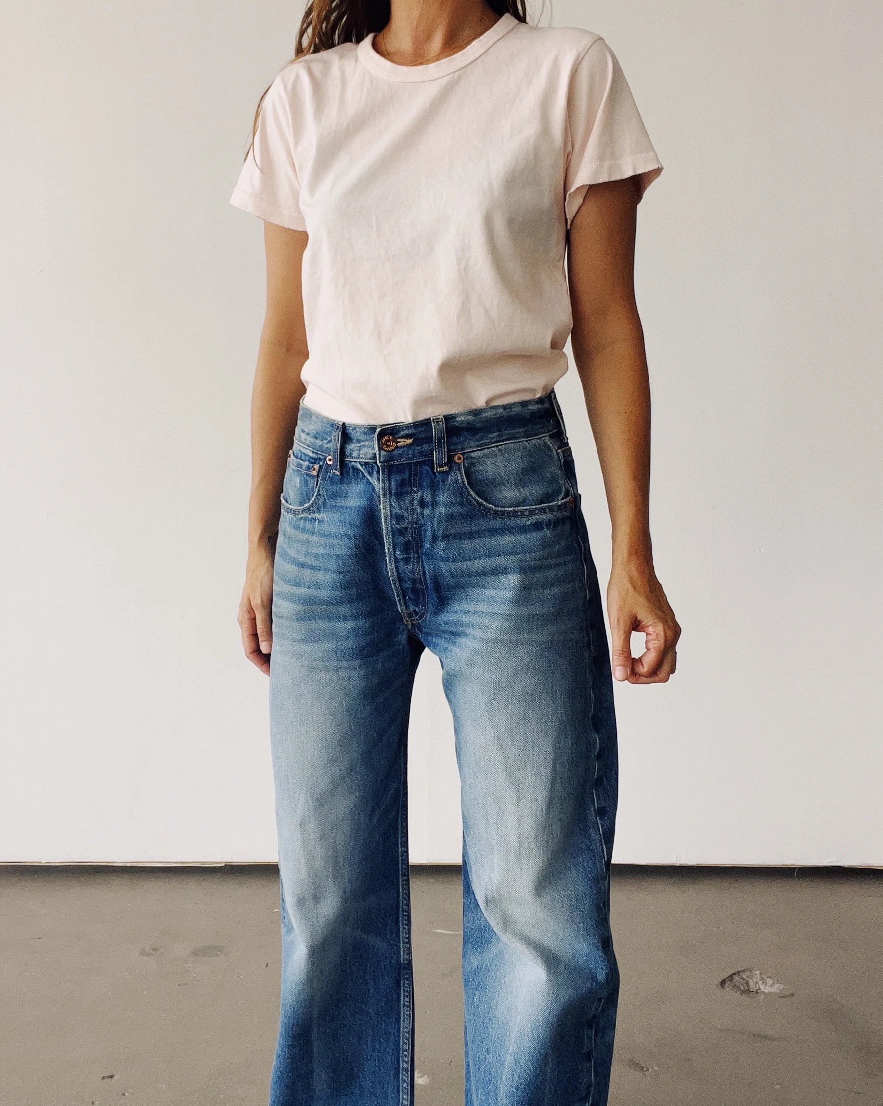 A woman, face not visible, stands against a plain background wearing Hey Gang's The Crew 26 light pink 100% cotton short-sleeve t-shirt with a vintage fit, tucked into high-waisted wide-leg blue jeans.