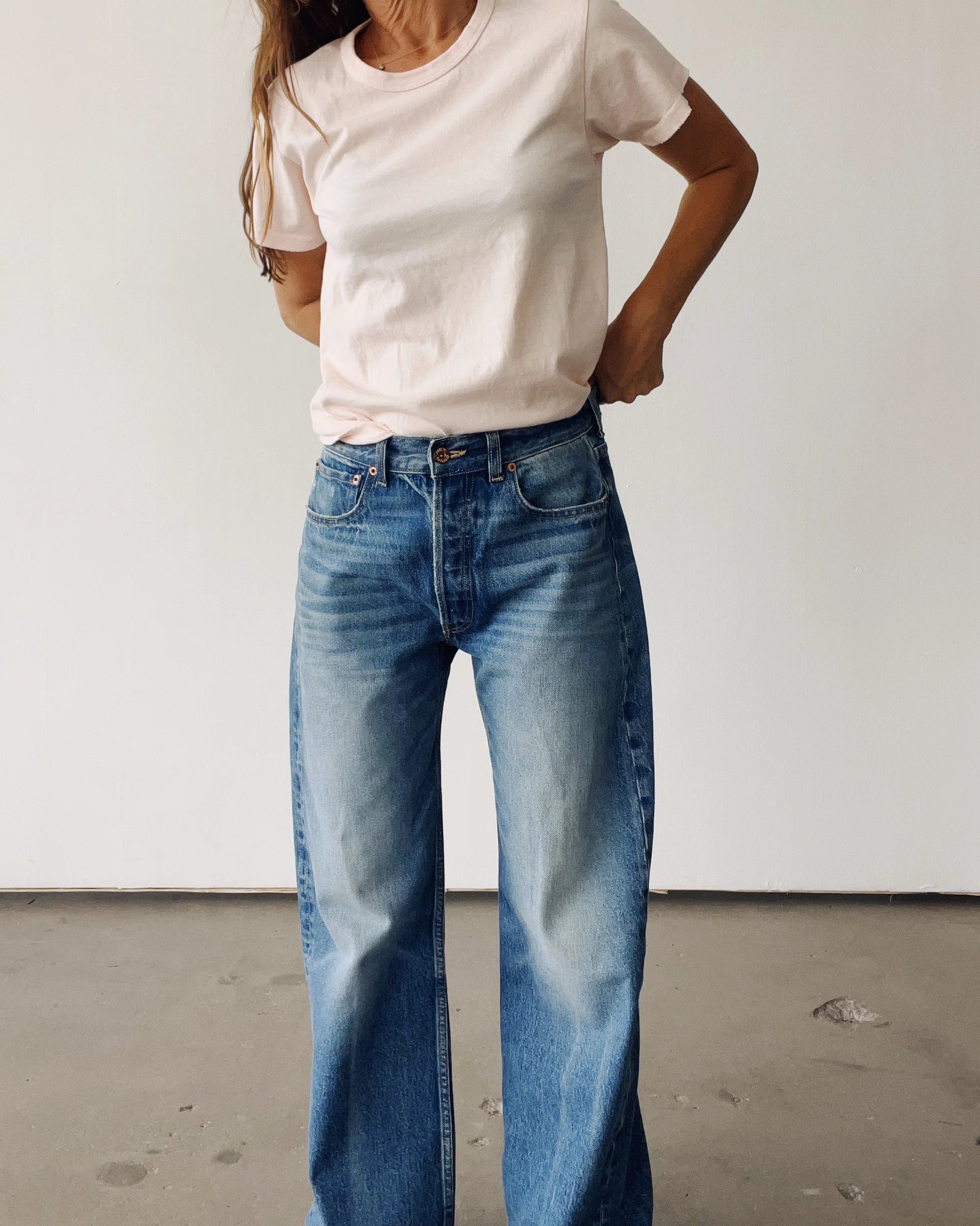 A person stands indoors against a plain wall, wearing Hey Gang’s The Crew 26 light pink short-sleeve t-shirt and vintage fit, high-waisted loose blue jeans with hands on hips; their head is partially out of frame.