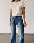 A woman, face not visible, stands against a plain background wearing Hey Gang's The Crew 26 light pink 100% cotton short-sleeve t-shirt with a vintage fit, tucked into high-waisted wide-leg blue jeans.